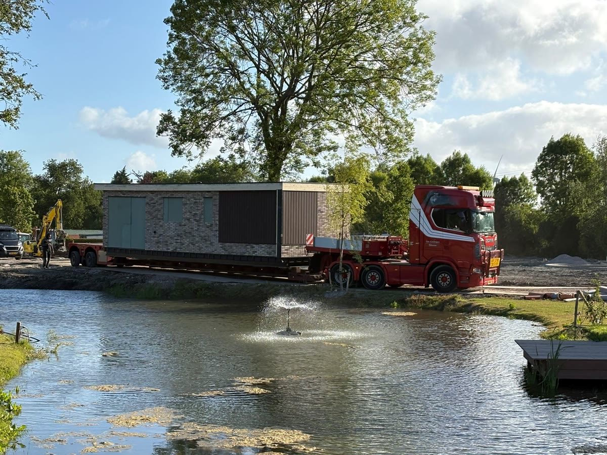 Rode truck van Van Lambalgen met een chalet op de oplegger langs een vijver op een vakantiepark