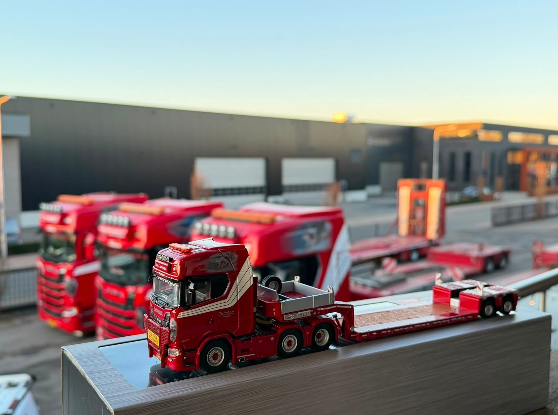 Miniatuur van de rode Van Lambalgen Scania met dieplader, met de echte trucks op de achtergrond
