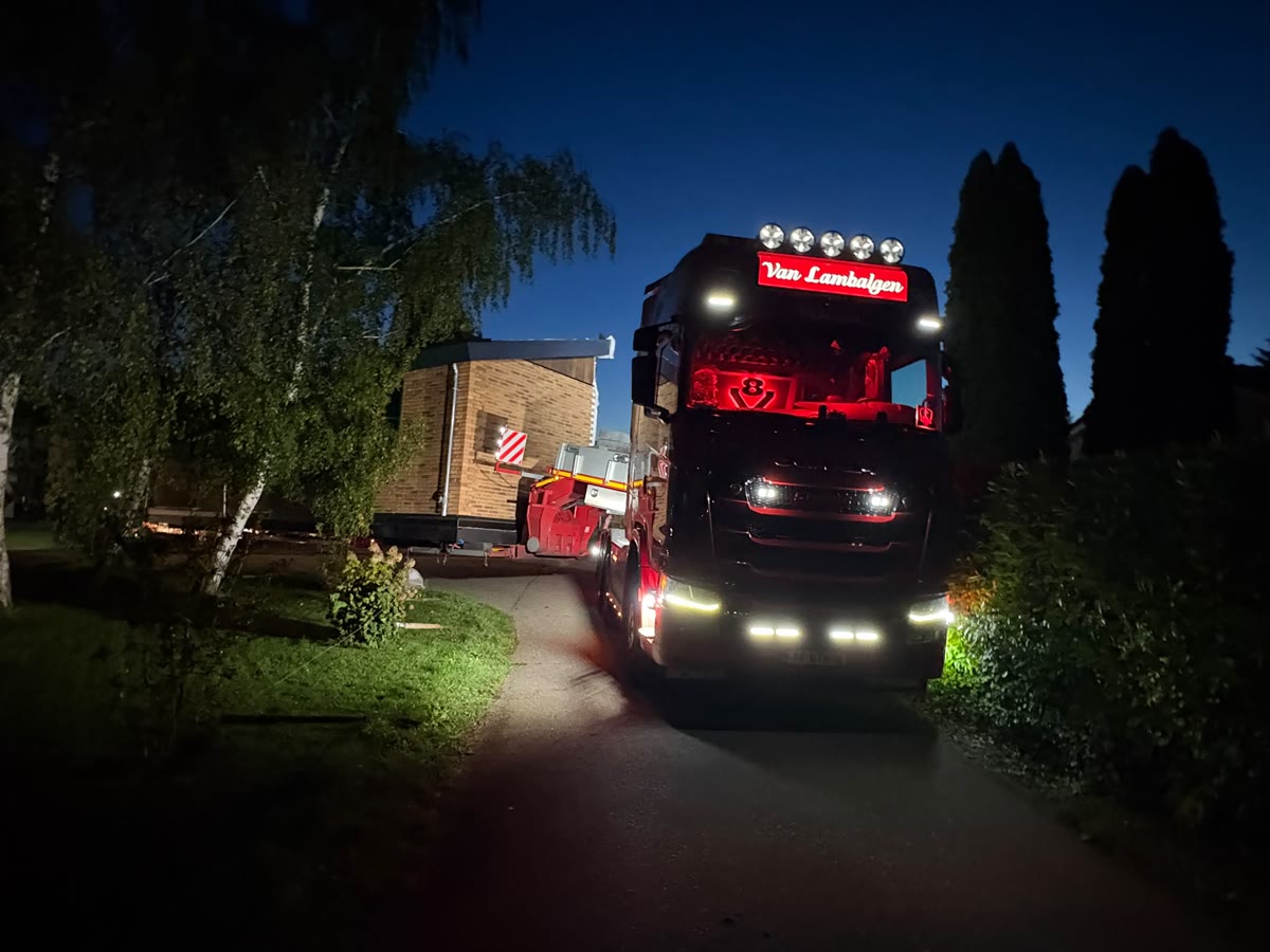 Truck met verlichte naam Van Lambalgen rijdt 's avonds met een woning over een smalle weg