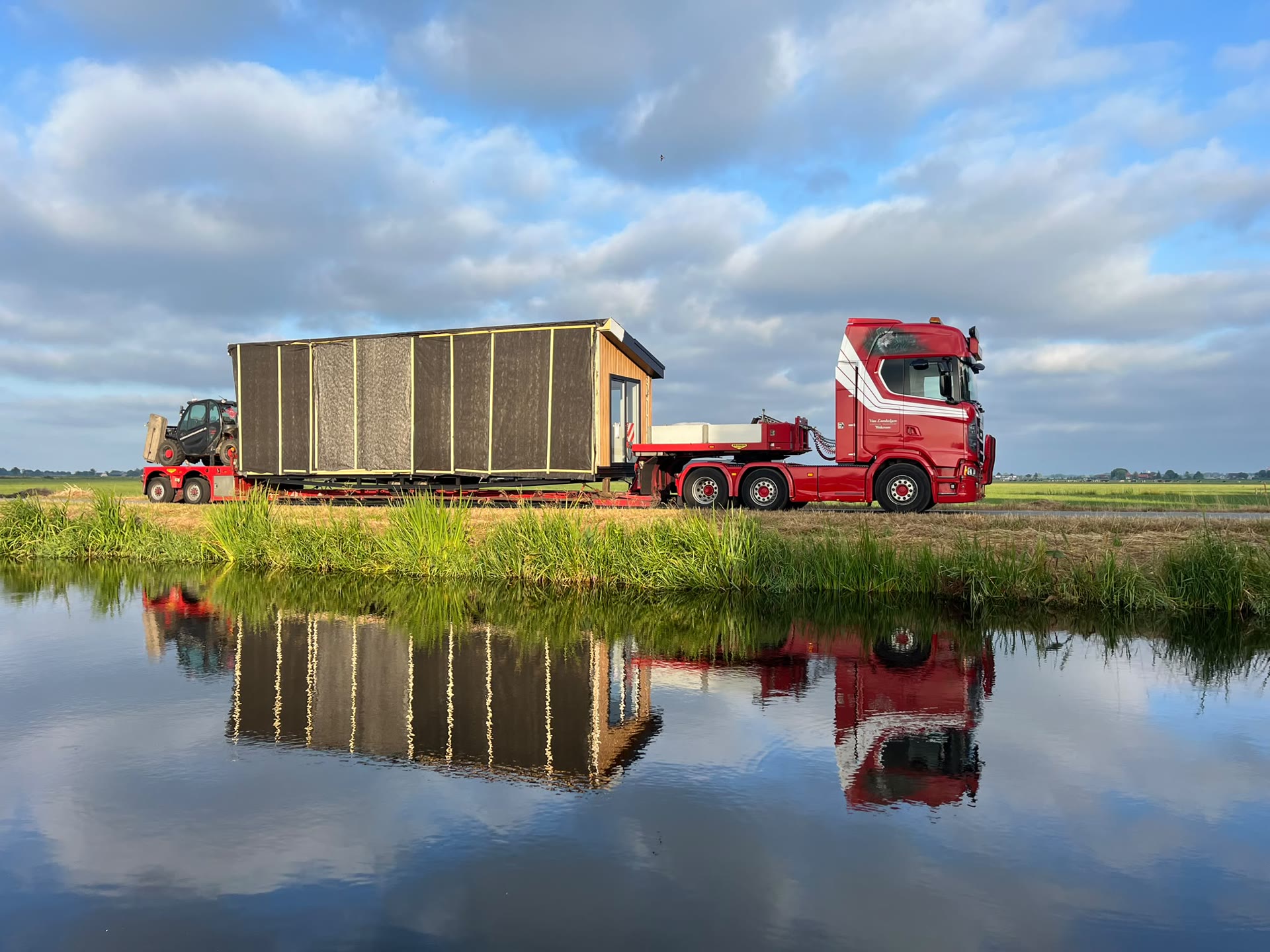 Truck met een houten woning weerspiegeld in een sloot in de polder